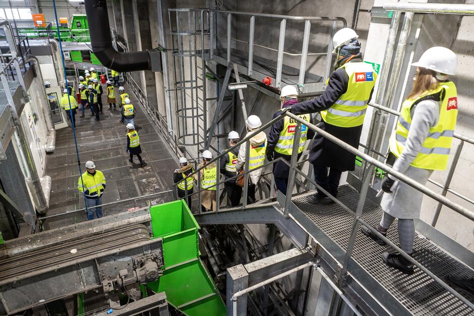 Grupa ludzi w kaskach ochronnych i kamizelkach zwiedzająca zakład