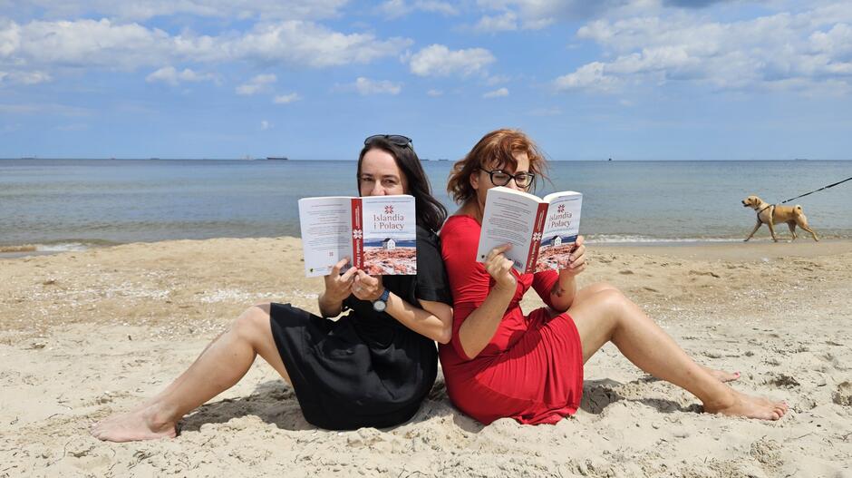 dwie kobiety siedzą na plaży plecami do siebie, trzymają książki, których okładki pokazują