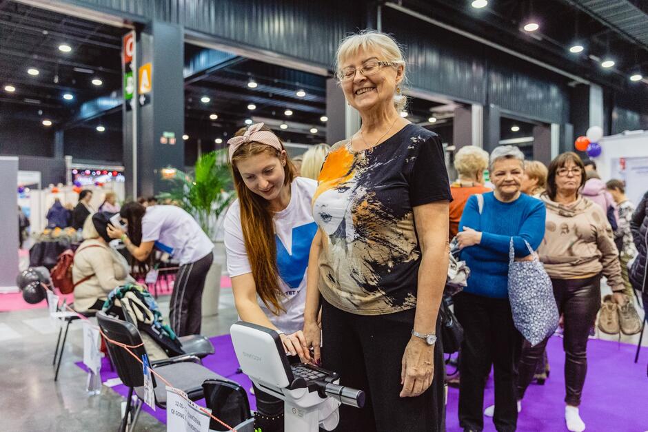 na zdjęciu starsza kobieta stoi na urządzeniu, które przypomina wagę, obok niej stoi młoda kobieta