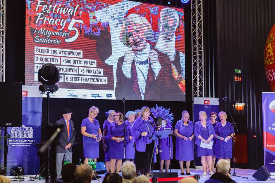 na scenie stoi grupa starszych kobiet ubrana w niebieskie garsonki, w tle duży ekran z wyświetlaną informacją na temat oferty targów