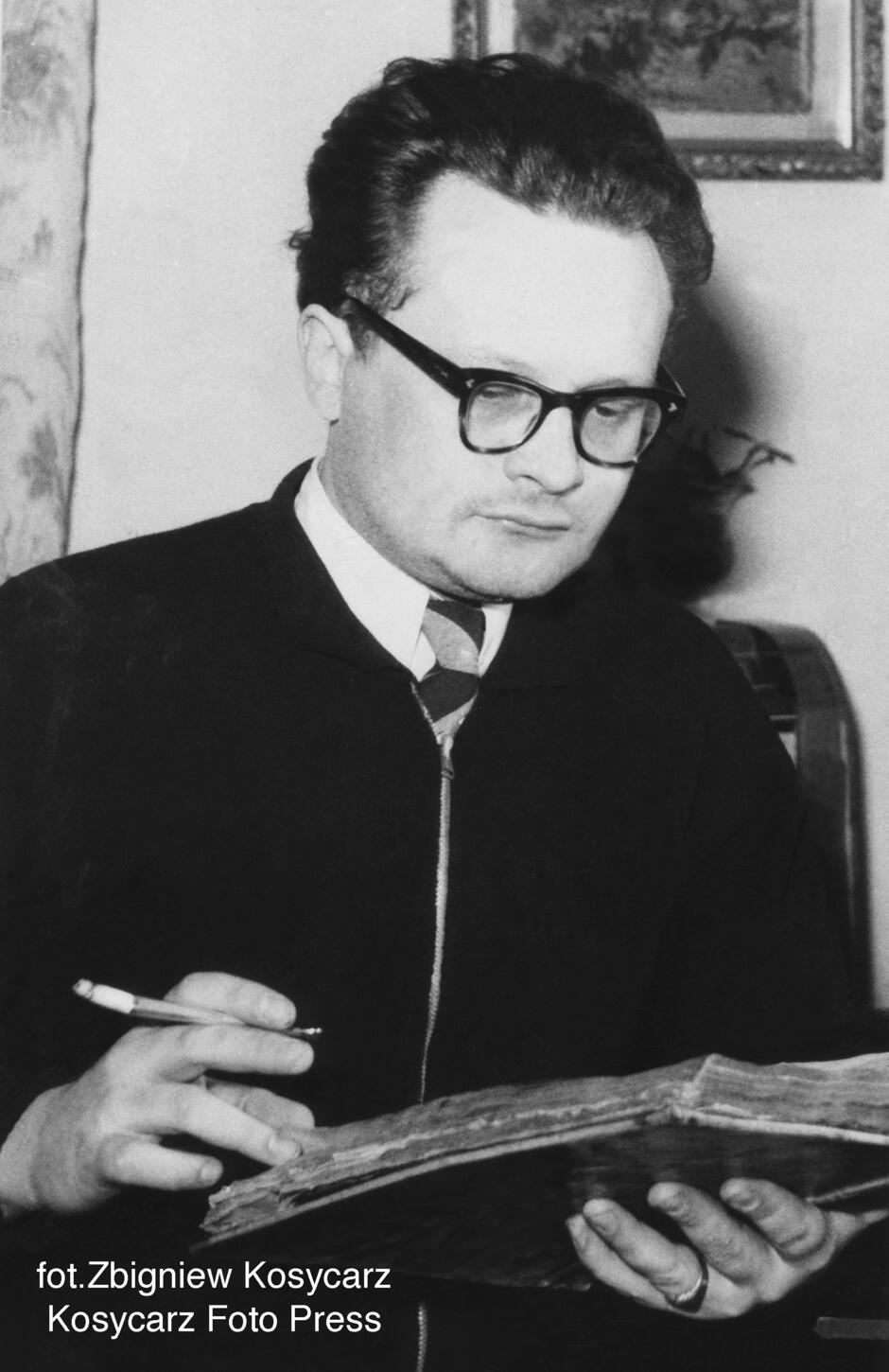 czarno-białe zdjęcie młodego mężczyzny w okularach pochylającego się nad książką z papierosem w ręce