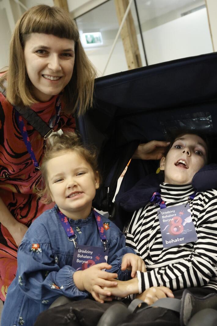niepełnosprawny leżący nastolatek w wózku, obok stoi jego mama i młodsza, ok 4-letnia siostra 