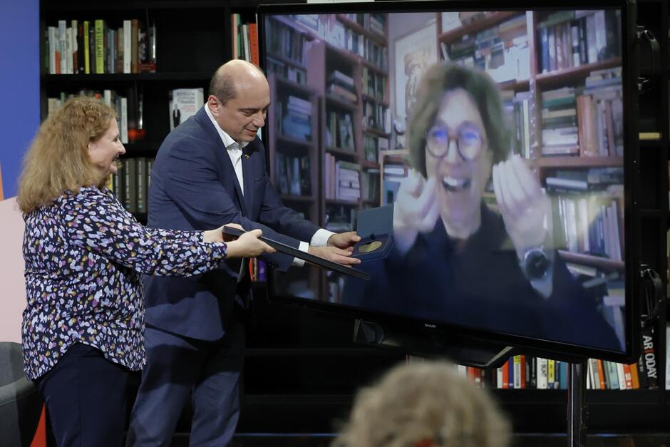 Kobieta i mężczyzna zbliżają do ekranu dyplom i medal, a na telewizorze jest uśmiechnięta kobieta