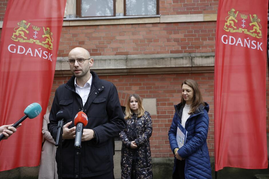 Łysawy mężczyzna w średnim wieku stoi w kurtce przed mikrofonami. Po bokach czerwone flagi z logo Gdańska, z tyłu trzy inne osoby