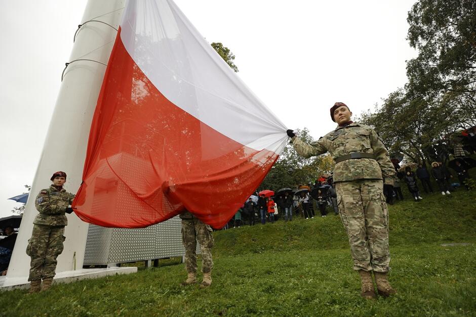 wielki maszt, po obu stronach stoją młode kobiety w mundurach, jedna przytrzymuje flagę RP jadącą na górę