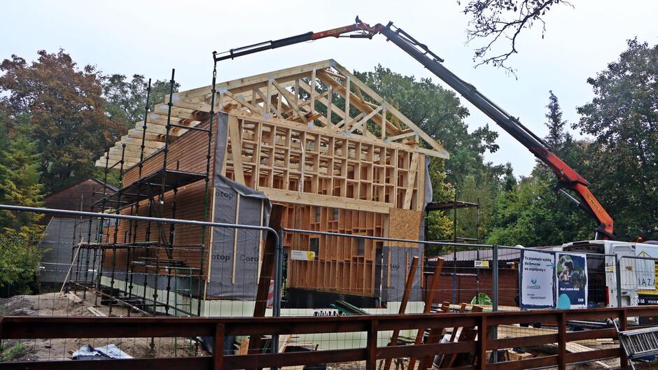 Na zdjęciu widać, jak żuraw podnosi kawałek drewna na dach domu, który jest w budowie. Drewno jest prawdopodobnie elementem konstrukcji dachu, który zostanie wykorzystany do podparcia lub wzmocnienia