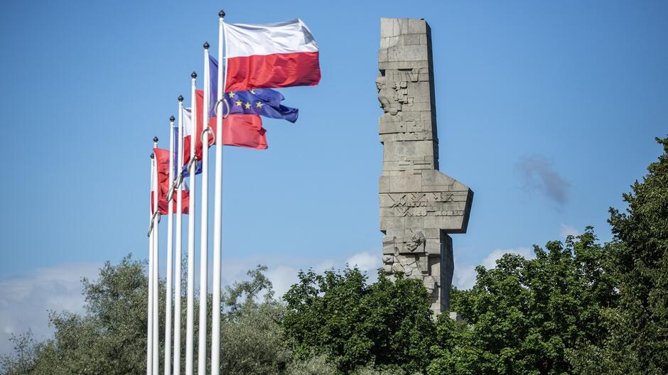 Na zdjęciu widać szereg flag powiewających przed pomnikiem. Flagi są białe, czerwone i niebieskie, co są kolorami narodowymi Polski. Pomnik jest dużym, kamiennym monumentem, który przedstawia żołnierzy polskich walczących w obronie Westerplatte w 1939 roku