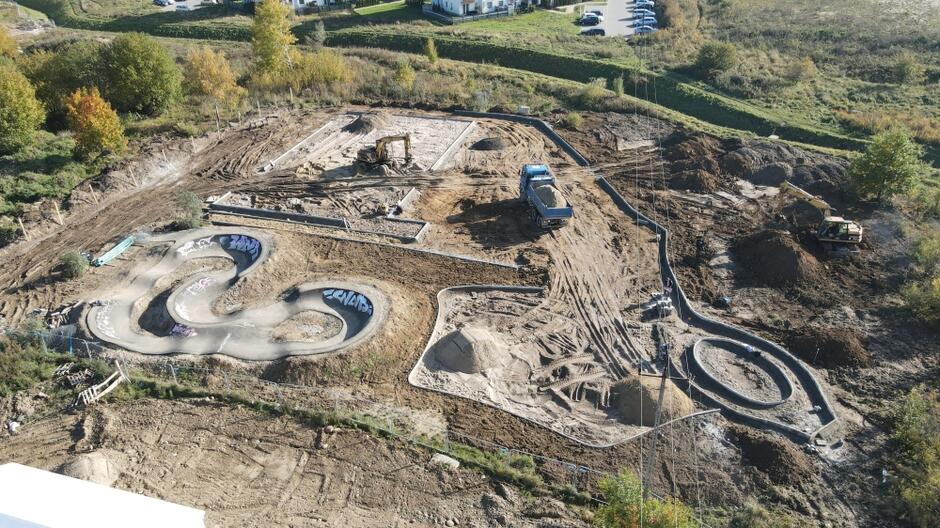 zdjęcie z drona, widać plac budowy, żółtą koparkę i rozkopany teren gruntowy