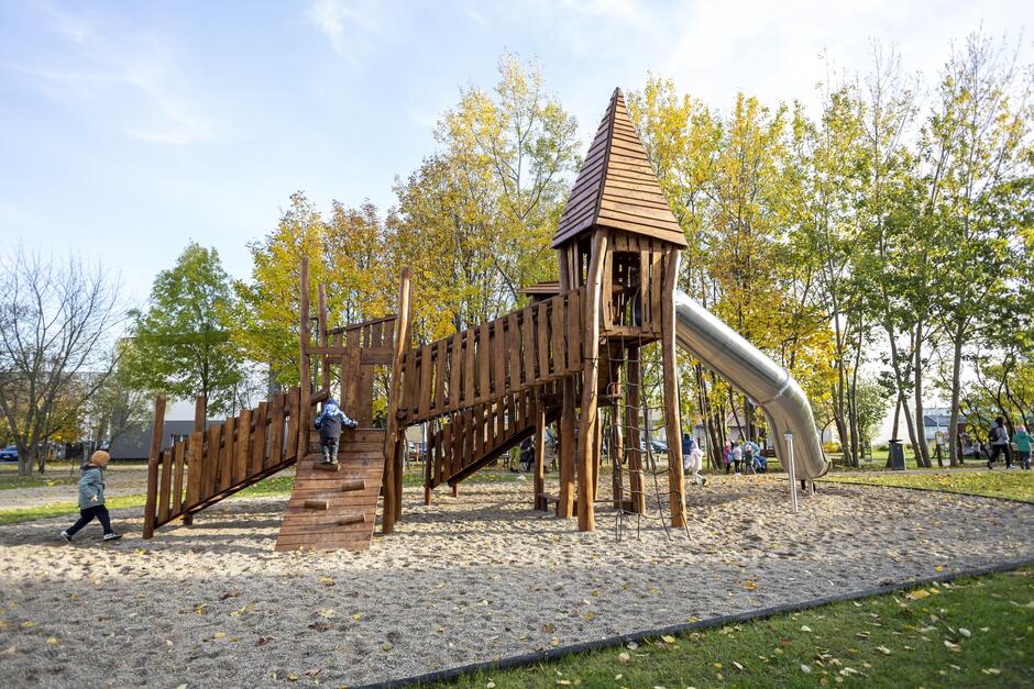 na zdjęciu drewniana konstrukcja przypominająca zamek z metalową rurą która pełni rolę zjeżdżalni, w tle jesienne drzewa