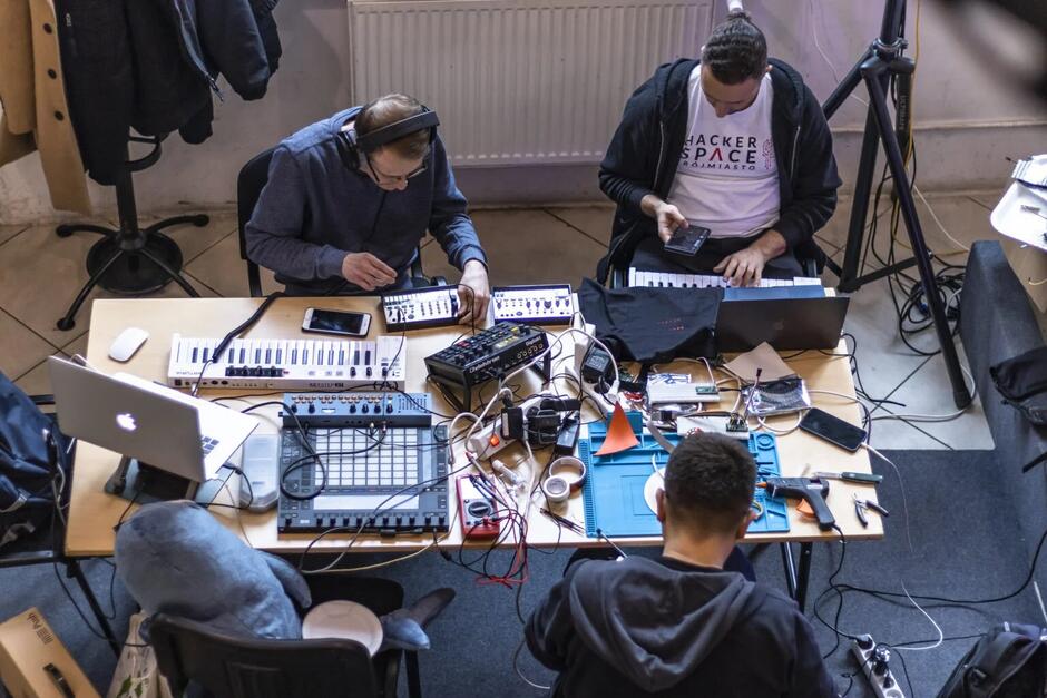 stół zastawiony laptopami i elektronicznymi instrumentami klawiszowymi, przy nim trzech mężczyzn