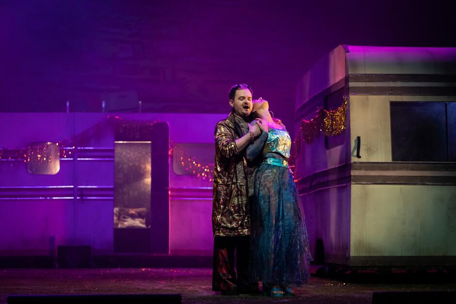 dwoje aktorów w miłosnym uścisku na scenie, w tle przyczepa kempingowa