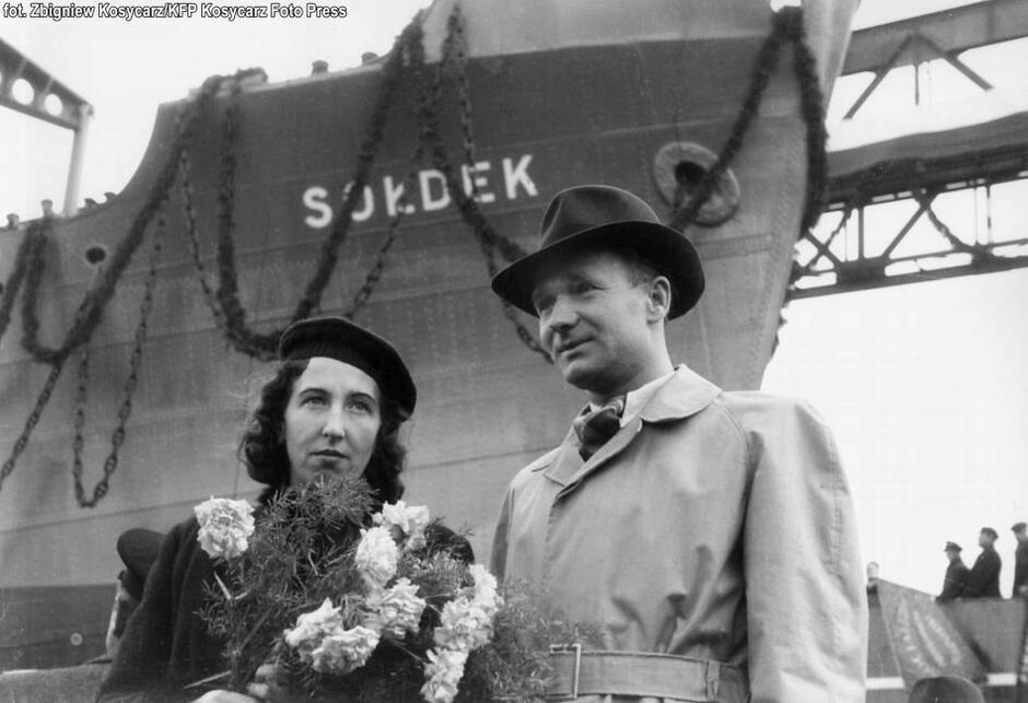 czarno-białe zdjęcie kobiety z kwiatami i mężczyzny w kapeluszu, oboje w jesiennych płaszczach, w tle statek