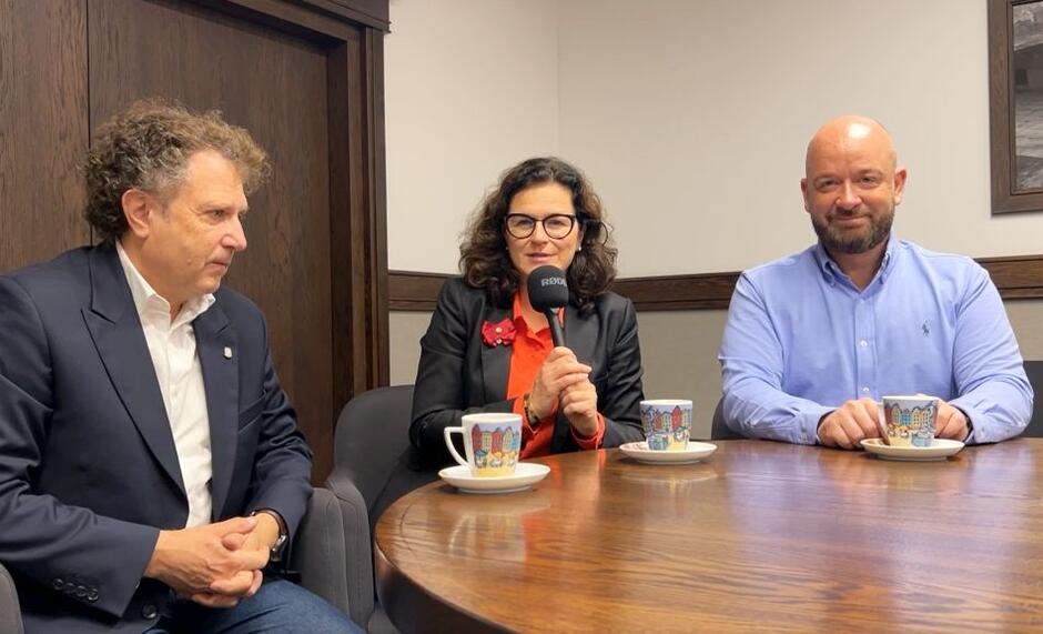 na zdjęciu dwóch mężczyzn w średnim wieku, i jedna kobieta siedząca między nimi, cała trójka siedzi za stołem, stoją przed nimi trzy kolorowe filiżanki