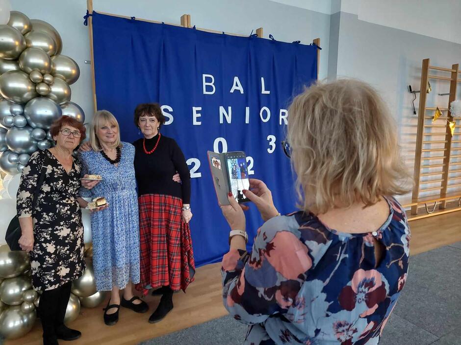 na zdjęciu starsza kobieta robi zdjęcie telefonem trzem innym starszym kobietom które pozują na niebieskim tle z napisem bal seniora