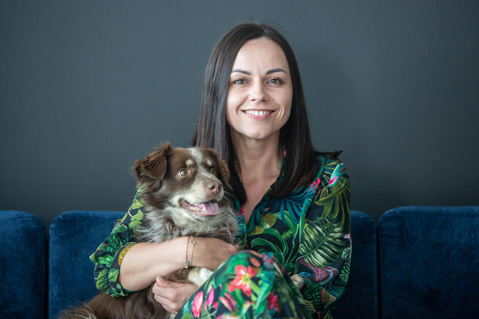 Brunetka w kolorowej sukience siedzi na granatowej kanapie, na kolanach trzyma brązowego psa - kundelka