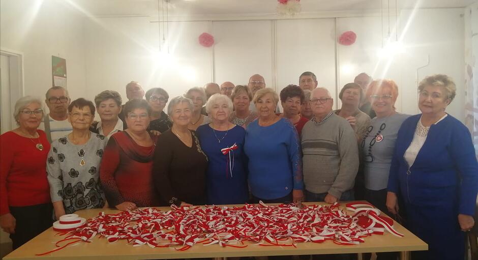grupa kilkudziesięciu kobiet i mężczyzn w starszym wieku stoi przed stołem, na którym leżą biało czerwone kokardy