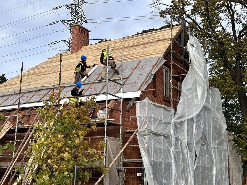 Na zdjęciu widać grupę mężczyzn stojących na dachu domu. Mężczyźni są ubrani w kombinezony robocze i kask. Niektórzy z nich trzymają w rękach narzędzia budowlane. Z tyłu zdjęcia widać dach innego domu
