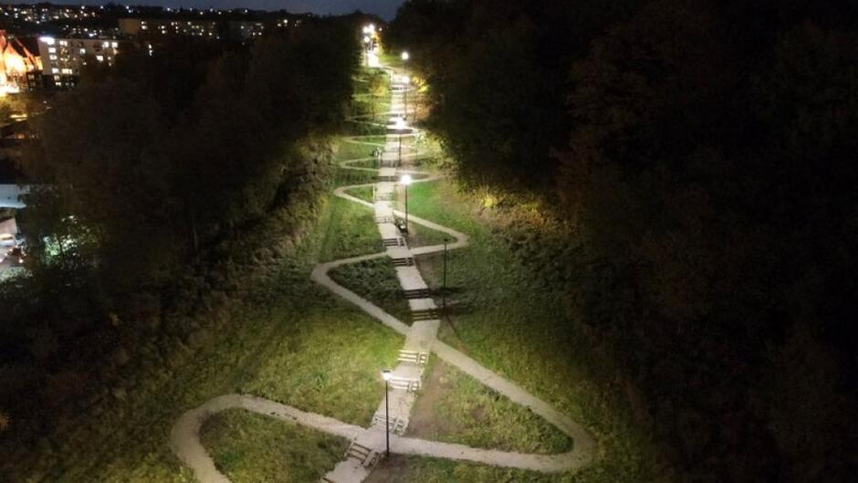 na zdjęciu świecą się latarnie, widać schody biegnące po zielonym wzgórzu i serpentynę chodnikową