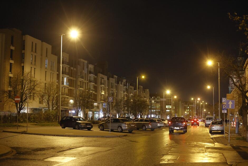 wieczorne zdjęcie ulicy długie ogrody, widać fragment ulicy, chodnika, zaparkowane samochody po lewej, w tle widać kilka niewysokich budynków