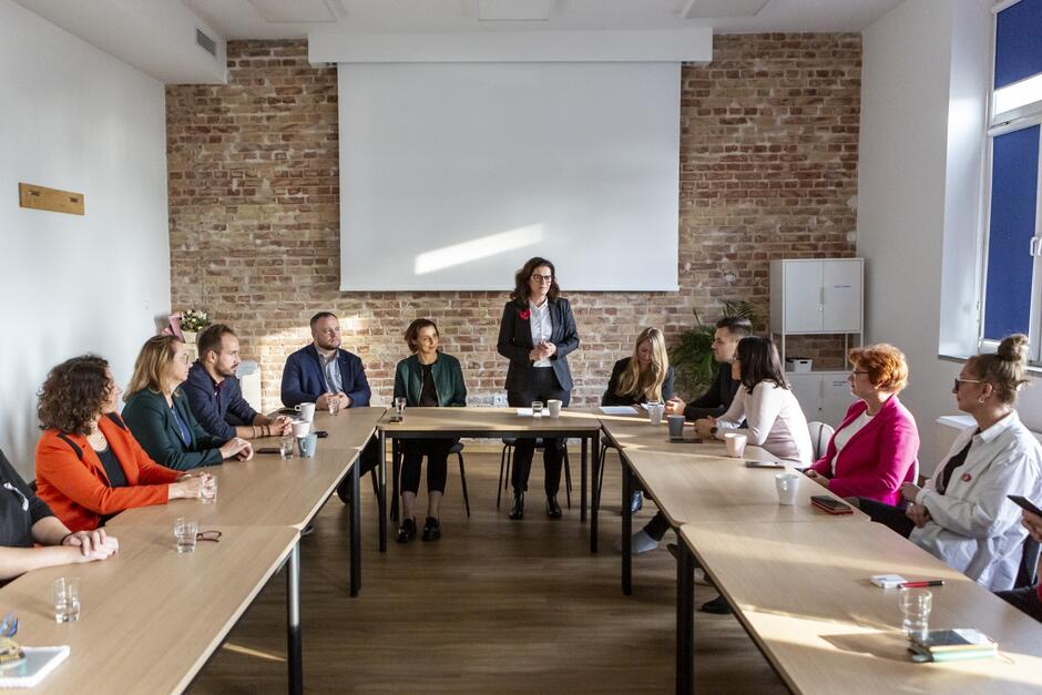 na zdjęciu około dziesięciu osób siedzi za stołem ustawionym w kształcie litery u, po środku jedna z osób w ciemnym kostiumie stoi i przemawia