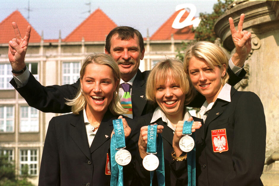 Trzy młode kobiety w ciemnych marynarkach z białym orłem na klapie trzymają w dłoniach medale. Za nimi mężczyzna z wąsem z rozpostartymi rękoma i palcami w kształcie litery V 