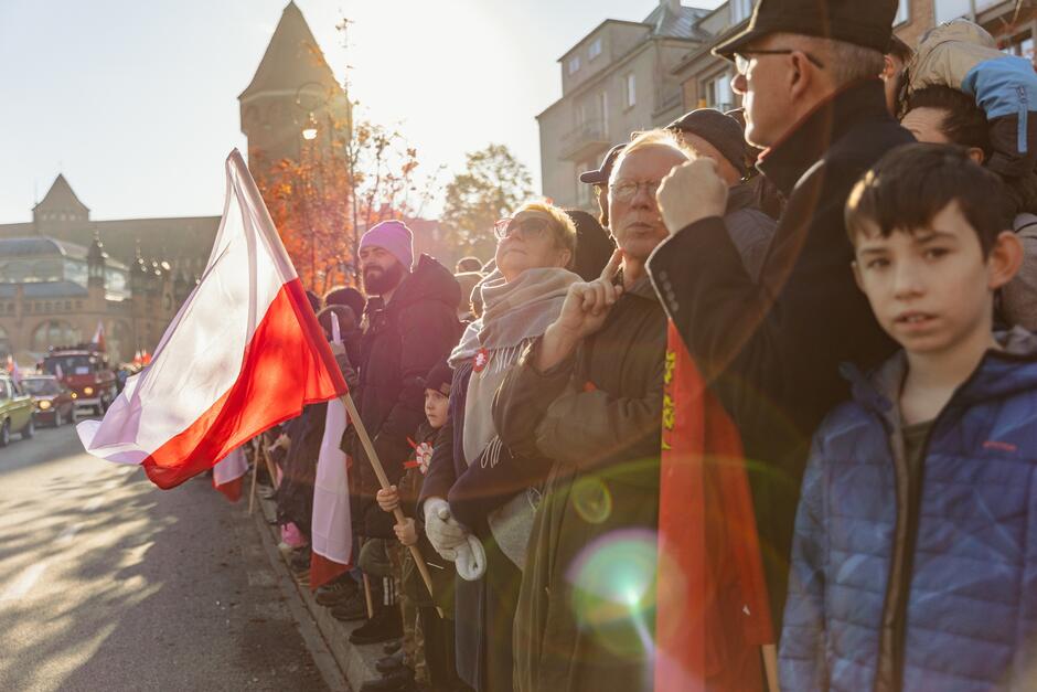 ludzie stoją na poboczu ulicy, widzowie parady, w dużej fladze Polski gra światło słońca