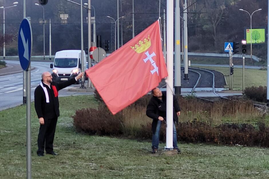 Na zdjęciu centralnie jest maszt z opuszczoną jeszcze - ale już przymocowaną do linki - flagą Gdańska, którą podtrzymuje mężczyzna. Mężczyzna stoi na prawo od masztu 