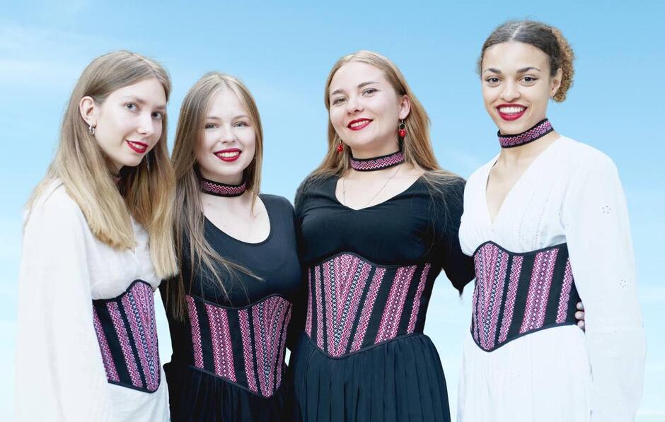 4 wokalistki w białych i czarnych sukienkach z kolorowym szerokim pasem obejmują się ramionami