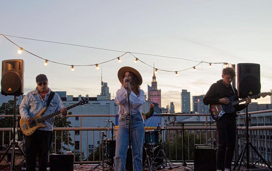 troje muzyków występuje na scenie na tle Pałacu Kultury i wieżowców w centrum Warszawy. Wokalistka w kapeluszu śpiewa, a dwaj muzycy grają na gitarach