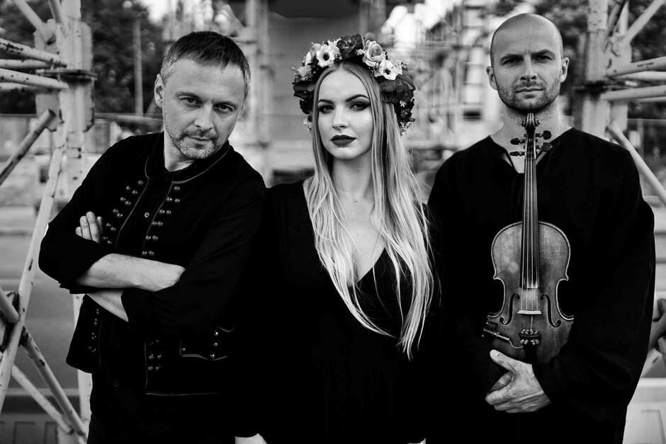  czarno-białe zdjęcie przedstawia członków zespołu. Pośrodku stoi wokalistka z wiankiem na głowie, po bokach muzycy, ten z prawej strony trzyma skrzypce