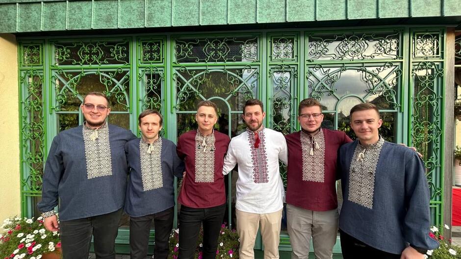 sześciu muzyków w wyszywanych koszulach stoi przed pięknie ozdobioną ornamentami zieloną bramą