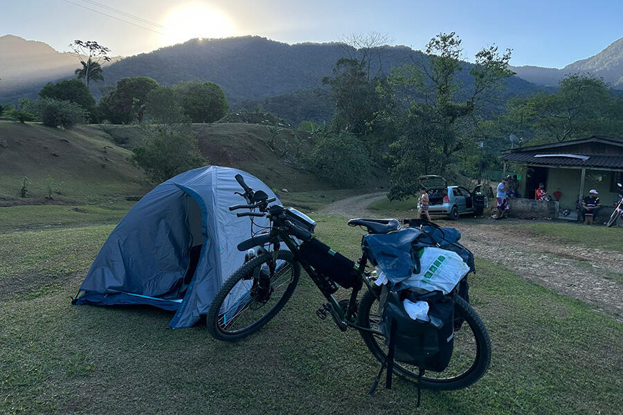 Na zdjęciu widać rower zaparkowany obok namiotu w polu. Rower jest czarny i składany, a namiot jest niebieski i zielony. Pole jest zielone i zadbane, a na niebie widać błękitne niebo