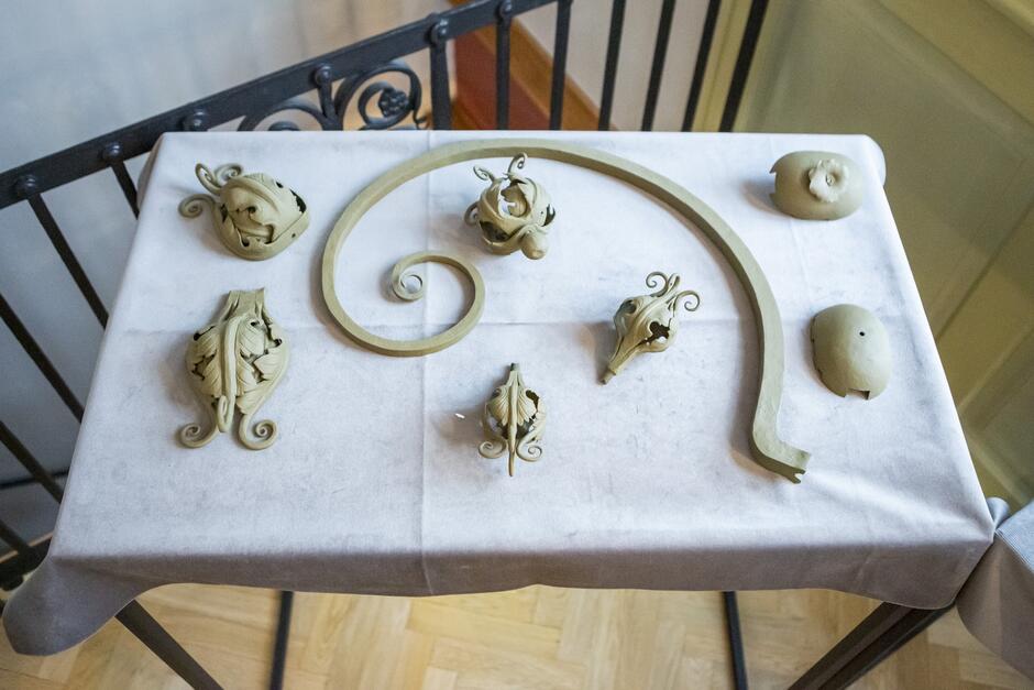Na zdjęciu widać stół z różnymi przedmiotami z kutego żelaza. Na stole znajduje się wazon, świecznik, lampa i kilka innych przedmiotów. Wszystkie przedmioty są wykonane z kutego żelaza i mają różne kształty i wzory. 