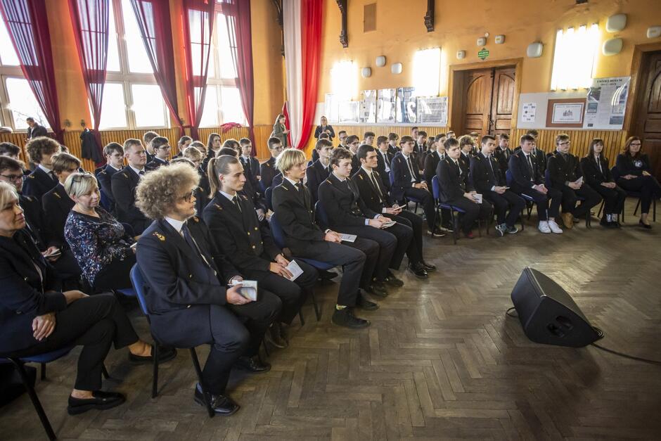 na zdjęciu grupa ponad pięćdziesięciu uczniów w oficjalnych strojach szkolnych, to ciemne mundury, wszyscy siedzą na krzesłach w przestronnej auli