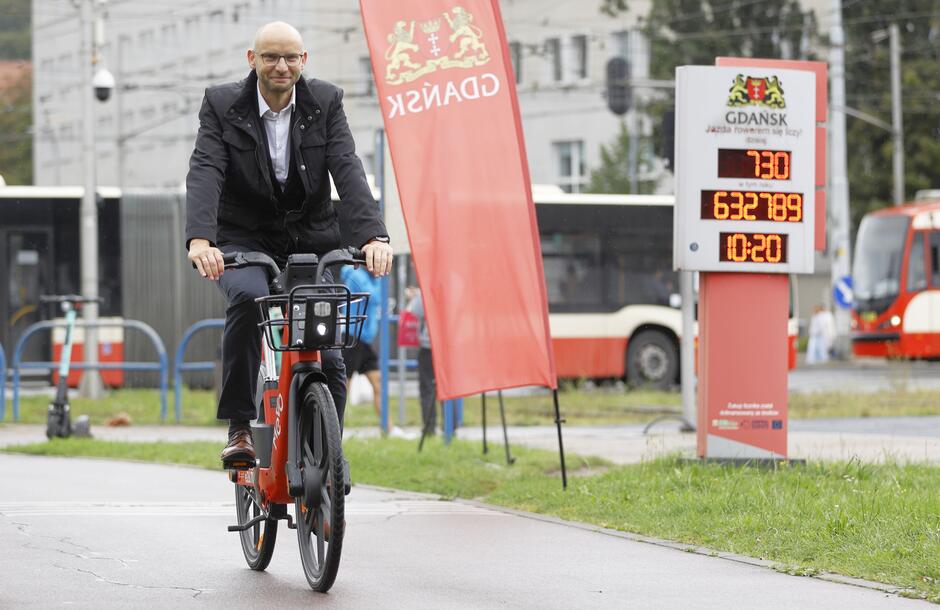 mężczyzna w średnim wieku, łysy w garniturze jedzie na czerwonym rowerze, w tle tramwaje i ulica