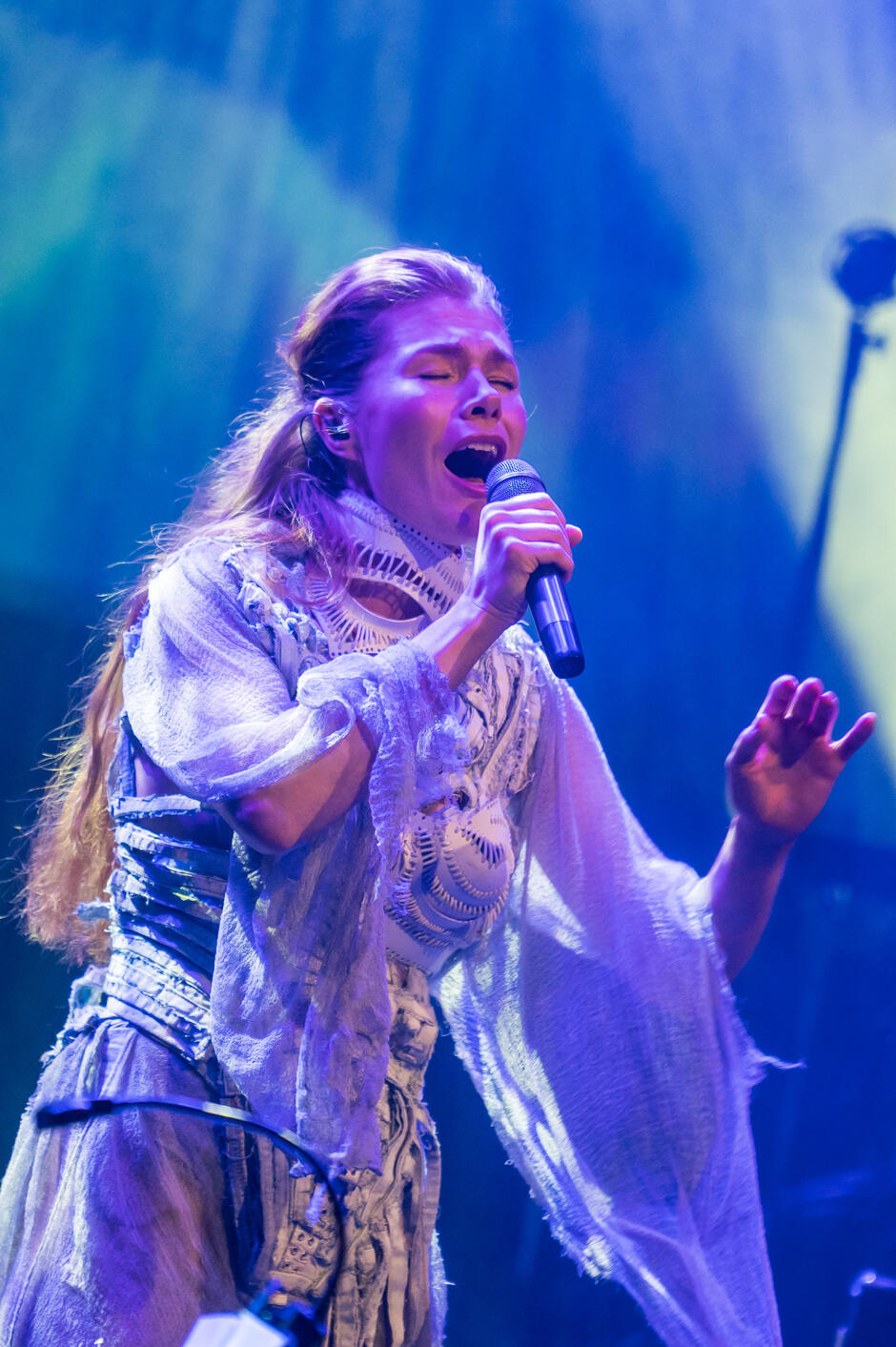 Młoda kobieta z długimi blond włosami, stoi na scenie, śpiewa do mikrofonu