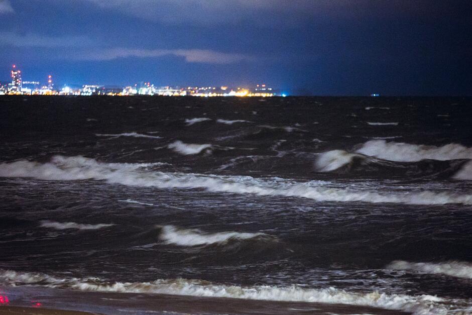 Na zdjęciu wzburzone morze nocą, w tle oświetlony pomost