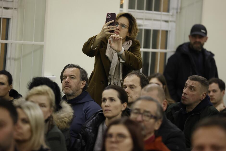 na zdjęciu kobieta w średnim wieku, w ciepłej kurtce, robi zdjęcie telefonem, pomiędzy nią widać kilka siedzących osób