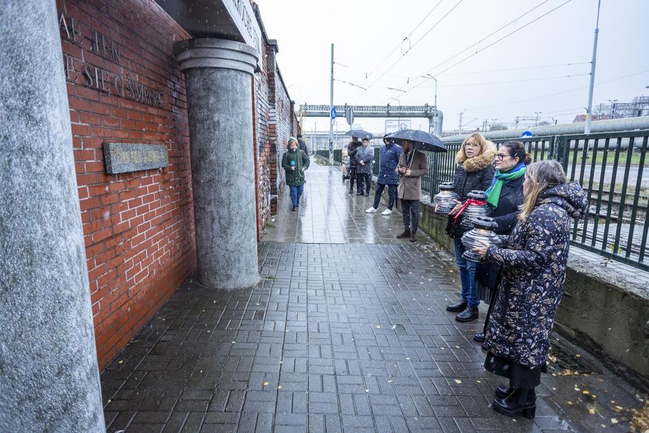 na zdjęciu po prawej stoją trzy kobiety w ciepłych kurtkach zimowych, trzymają znicze, w tle stoi kilka osób, po lewej widać fragment ogrodzenia