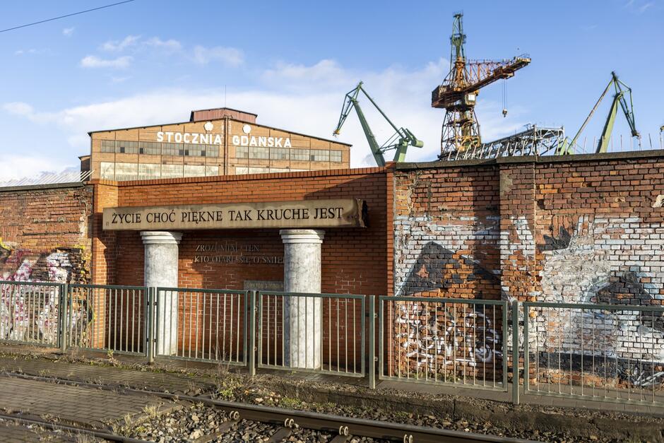 na zdjęciu mur, przy nim niewielki pomnik z kolumnami, w tle za murem widać halę i dźwigi stoczniowe