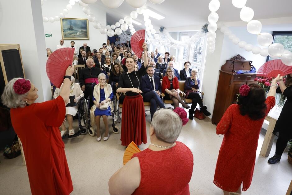 W głębi kobiety i mężczyźni w różnym wieku siedzą na krzesłach przed nimi starsze kobiety w czerwonych sukienkach tańczą z wachlarzami w dłoniach 