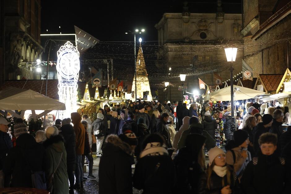 na zdjęciu kilka jarmarkowych stoisk, widać ozdoby wystawione na nich, i jedna osobę która stoi przy jednym ze stoisk