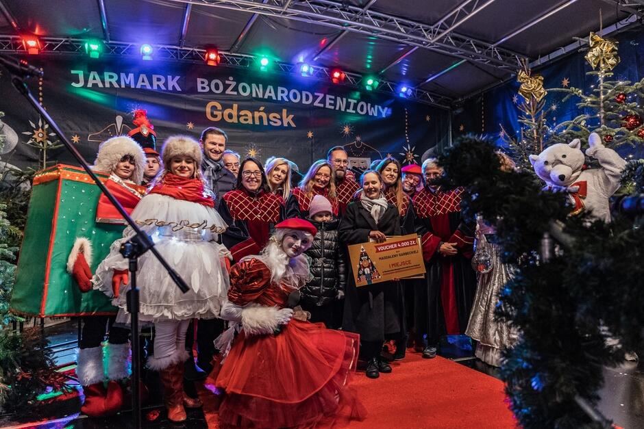 Scena, u góry napis Jarmark Bożenarodzeniowy Gdańsk. Pod napisem stoi kilkanaście osób, kobiet i mężczyzn, cześć z nich ma aksamitne peleryny, to Radni Miasta Gdańska. Obok nich młoda dziewczyna w białym, zimowym stroju, obok niej klęczy młoda dziewczyna ubrana na czerwono