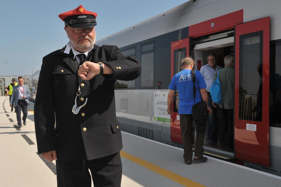Brodaty mężczyzna w okularach ubrany w mundur i czapkę konduktora pociągu, stoi na peronie przed pociągiem, spogląda na zegarek