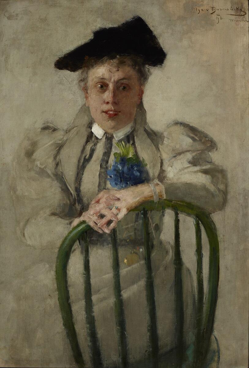 Portret: kobieta w jasnej sukni i ciemnym kapeluszu siedzi bokiem na krześle, opiera się ramionami o oparcie krzesła, które jest zrobione z drewnianych szczebelków. Splecione dłonie złożyła na oparciu. Uśmiecha się. Ma przypięty do sukni niebieski kwiat.  