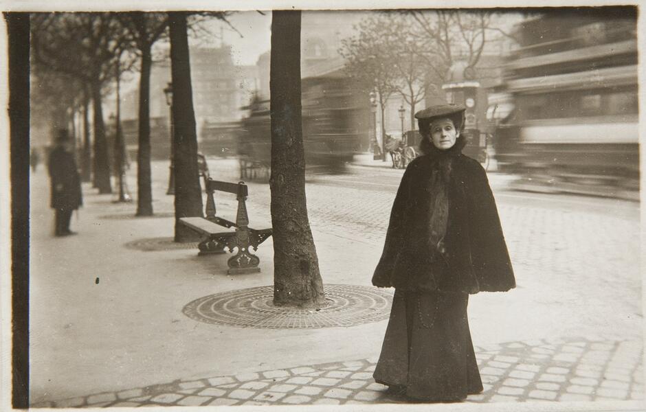 Na zdjęciu znajduje się kobieta w ciemnej długiej sukni, w pelerynie i kapeluszu. Stoi na ulicy, za nią znajdują się drzewa i ławka. Na końcu ulicy stoi mężczyzna w eleganckim stroju i kapeluszu.