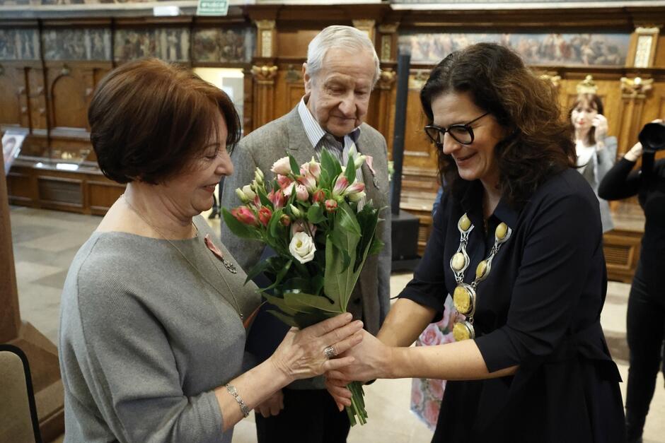 prezydent wręcza kobiecie kwiaty, obok stoi mężczyzna