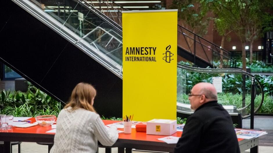 Kobieta i mężczyzna siedzą na ławce pod żółtym bannerem z napisem: Amnesty International. Za nimi widać zieleń i ruchome schody