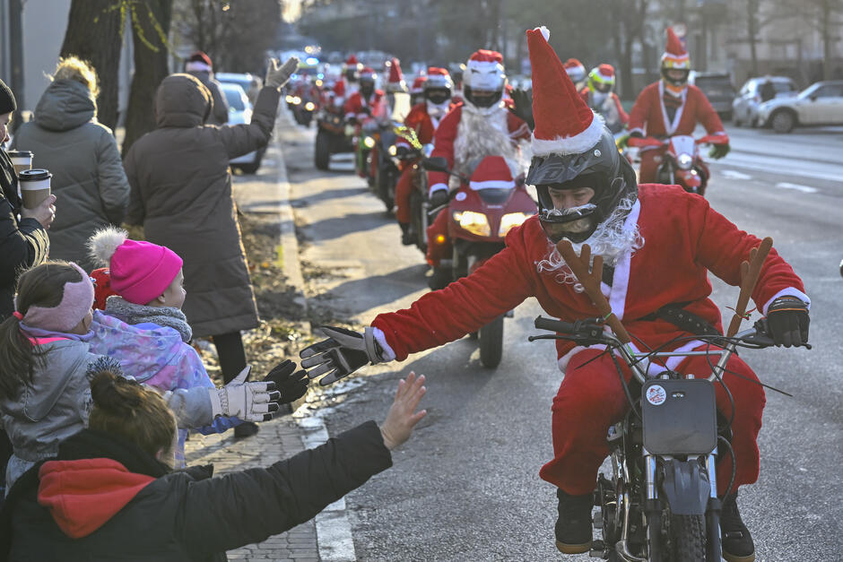 Mikołaje na motocyklach jadą ulicą. Ten na przedzie wychyla się i przybija piątkę z dziećmi stojącymi przy ulicy
