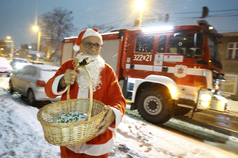 mężczyzna w stroju Mikołaja z koszykiem cukierków, w tle wóz strażacki